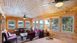 A cozy cabin interior with wooden walls, large windows, a dining area, and a seating area with a table and chairs. - Game Room, Dock Life, Fire Pit & Hot Tub! (Lake Ozark)