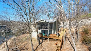 A cabin with a deck, surrounded by trees and a body of water. - Spacious, Central Location, Fire Pit, Boat Slip (Lake Ozark)