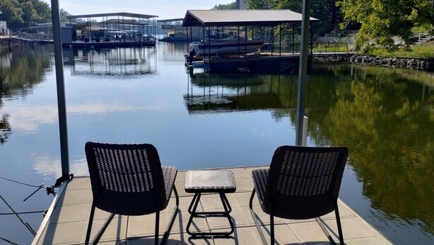 A covered outdoor seating area with two chairs and a small table, overlooking a calm lake with boats and buildings in the background. - Newly Updated in a Private Cove, With Firepit, Flat Lawn, Dock, Boat Slip & Kayaks in Osage Beach (Osage Beach)