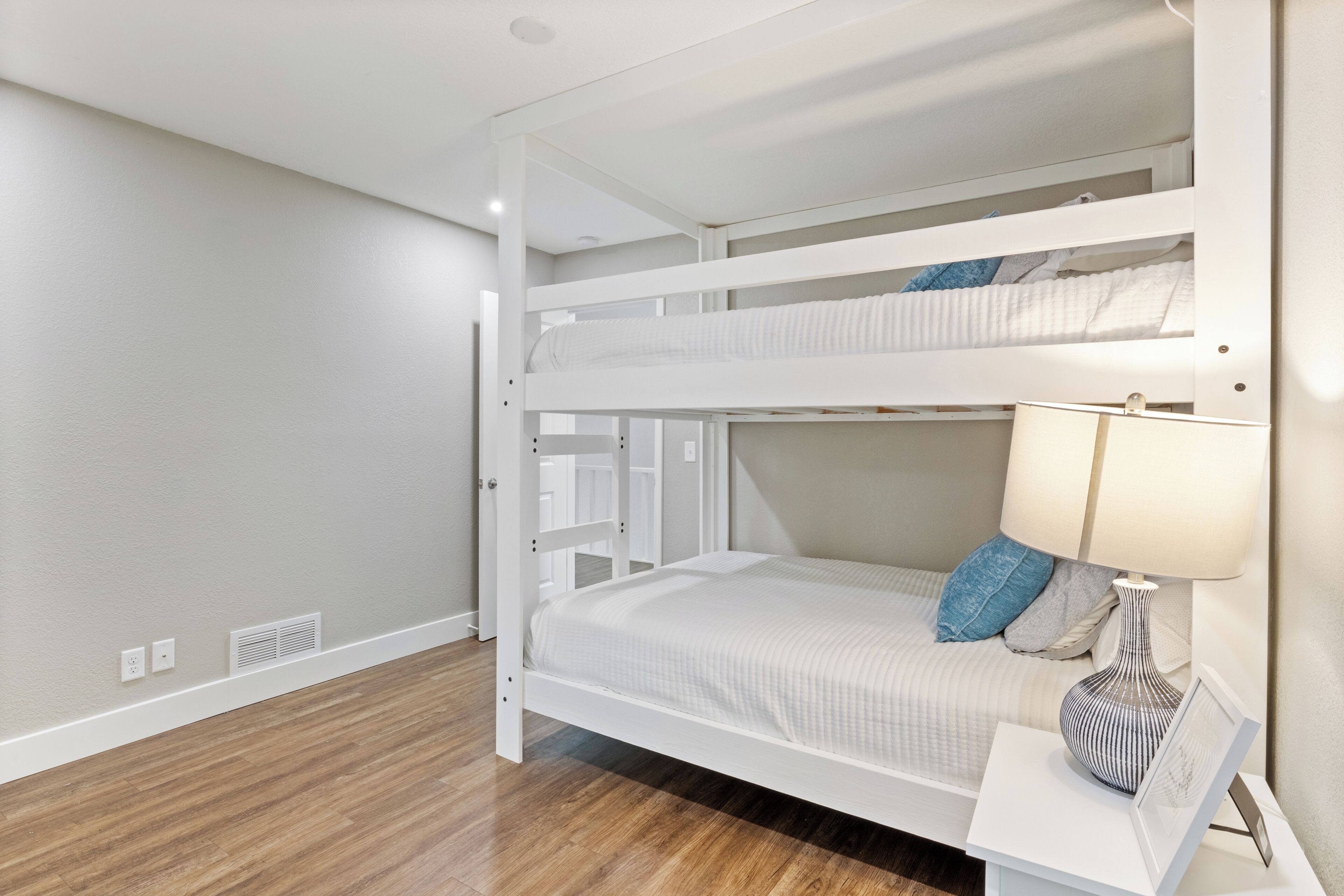 Habitación con literas de estructura blanca, una mesita de noche con lámpara y un marco de fotos.