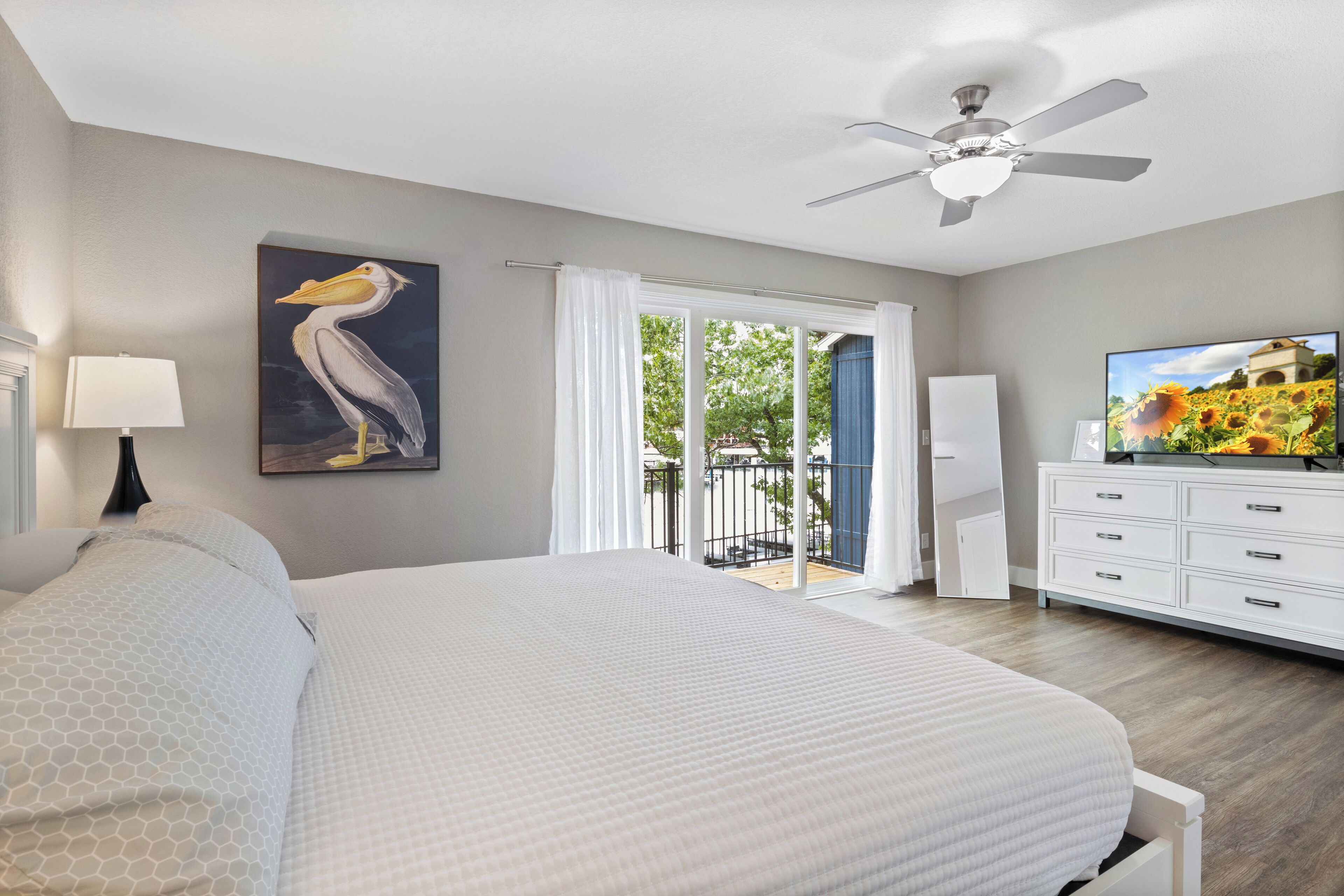 A bedroom with a large bed, a ceiling fan, a painting of a pelican, a television, and a view of the outdoors through a sliding door.