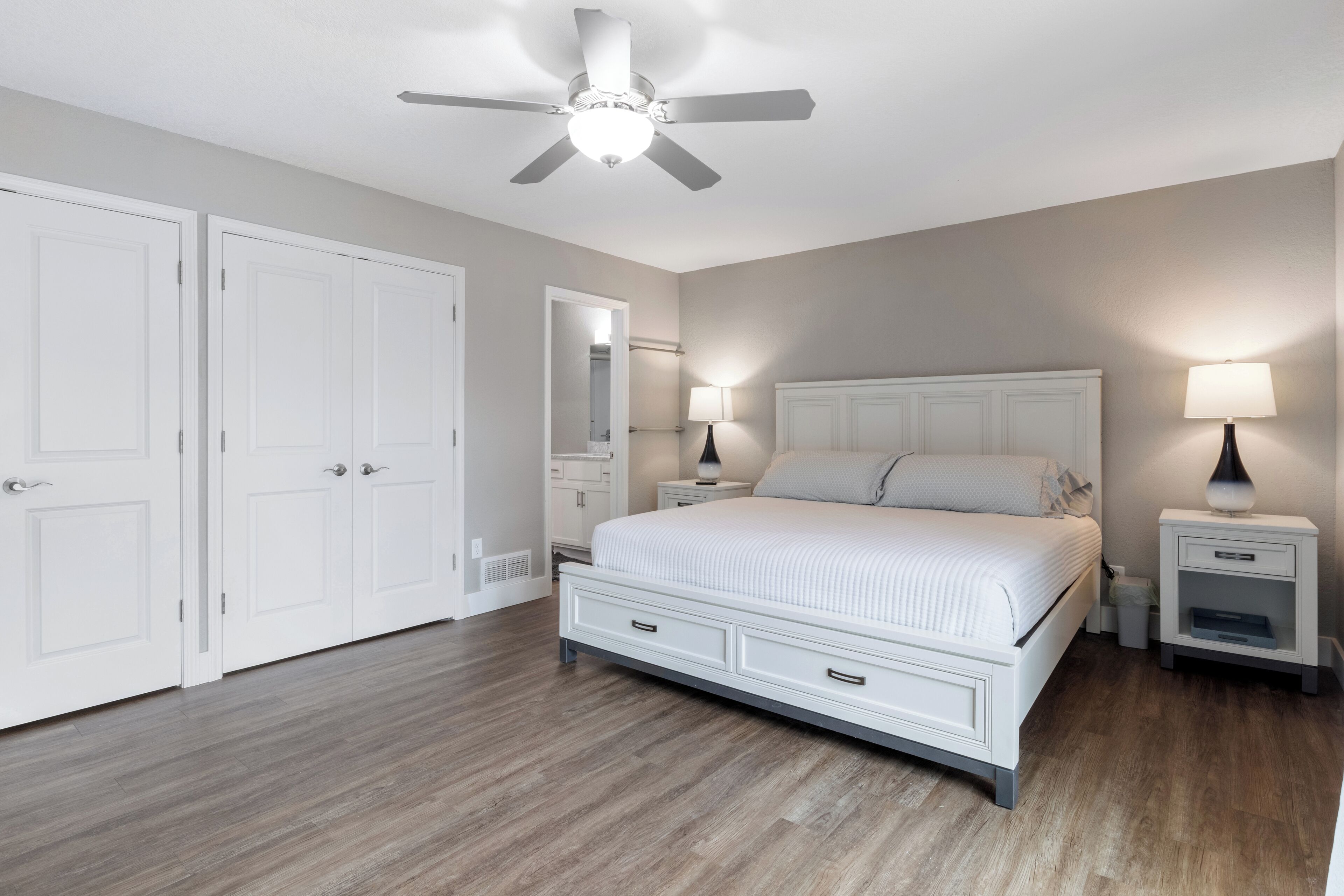 A bedroom with a large bed, white wardrobe, ceiling fan, bedside lamps, and a mirror.