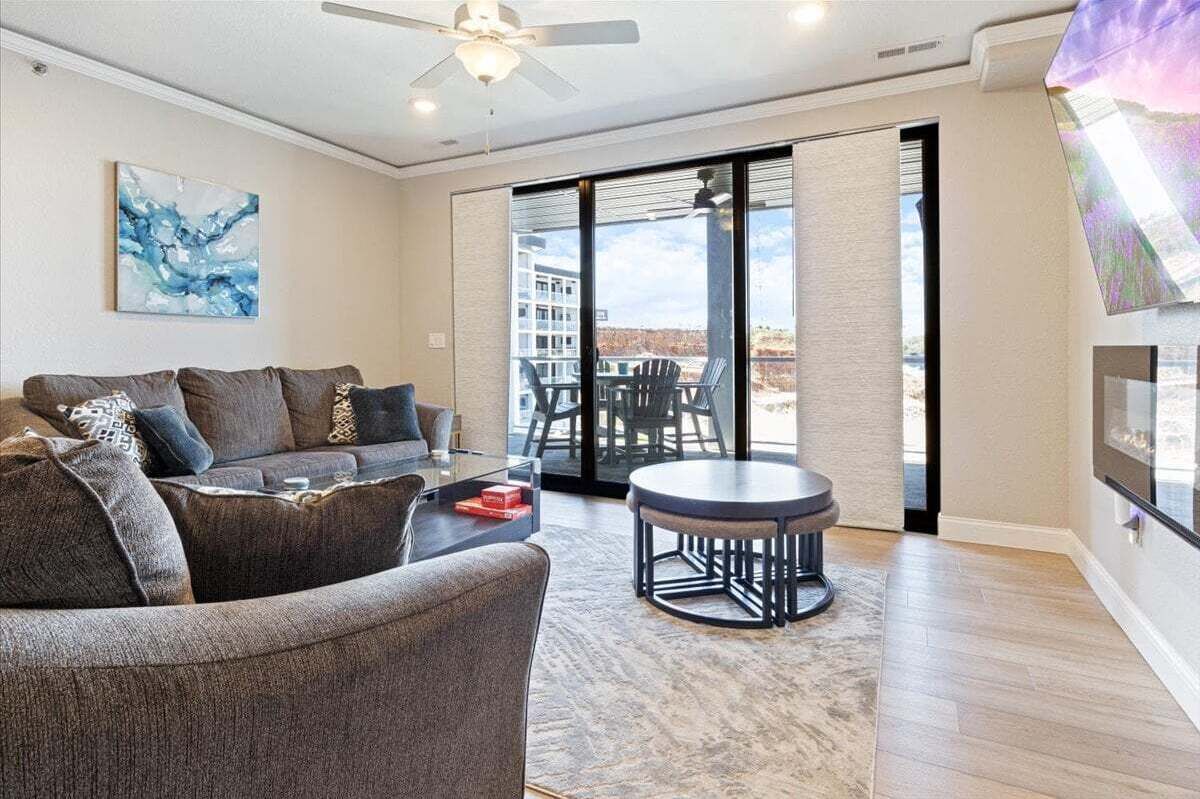 Uma sala de estar moderna com um sofá cinzento, uma mesa de centro e uma mesa redonda. Há uma janela grande com vista para edifícios e uma varanda.