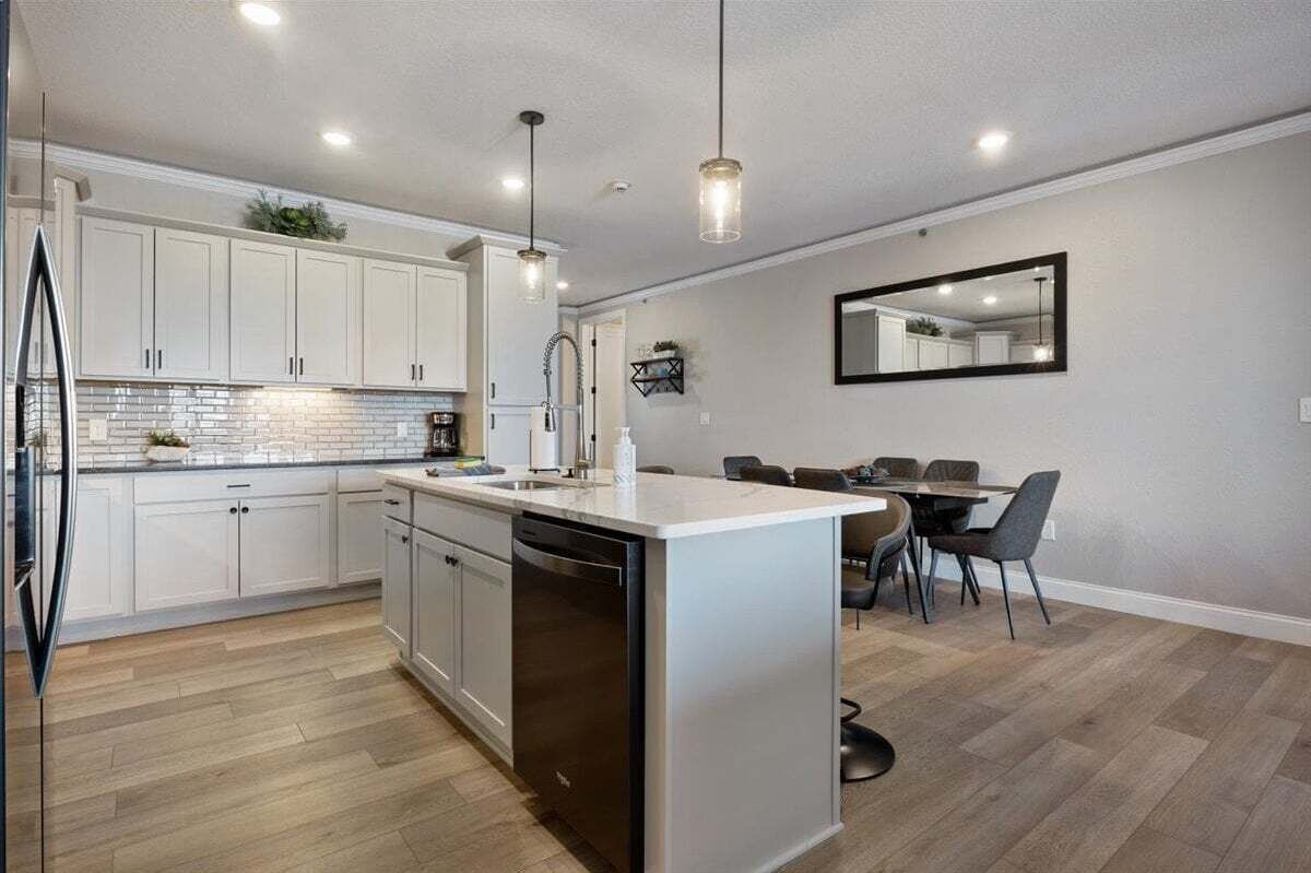 A modern kitchen with white cabinets, a dishwasher, and a central island. There is a dining area with a table and chairs.