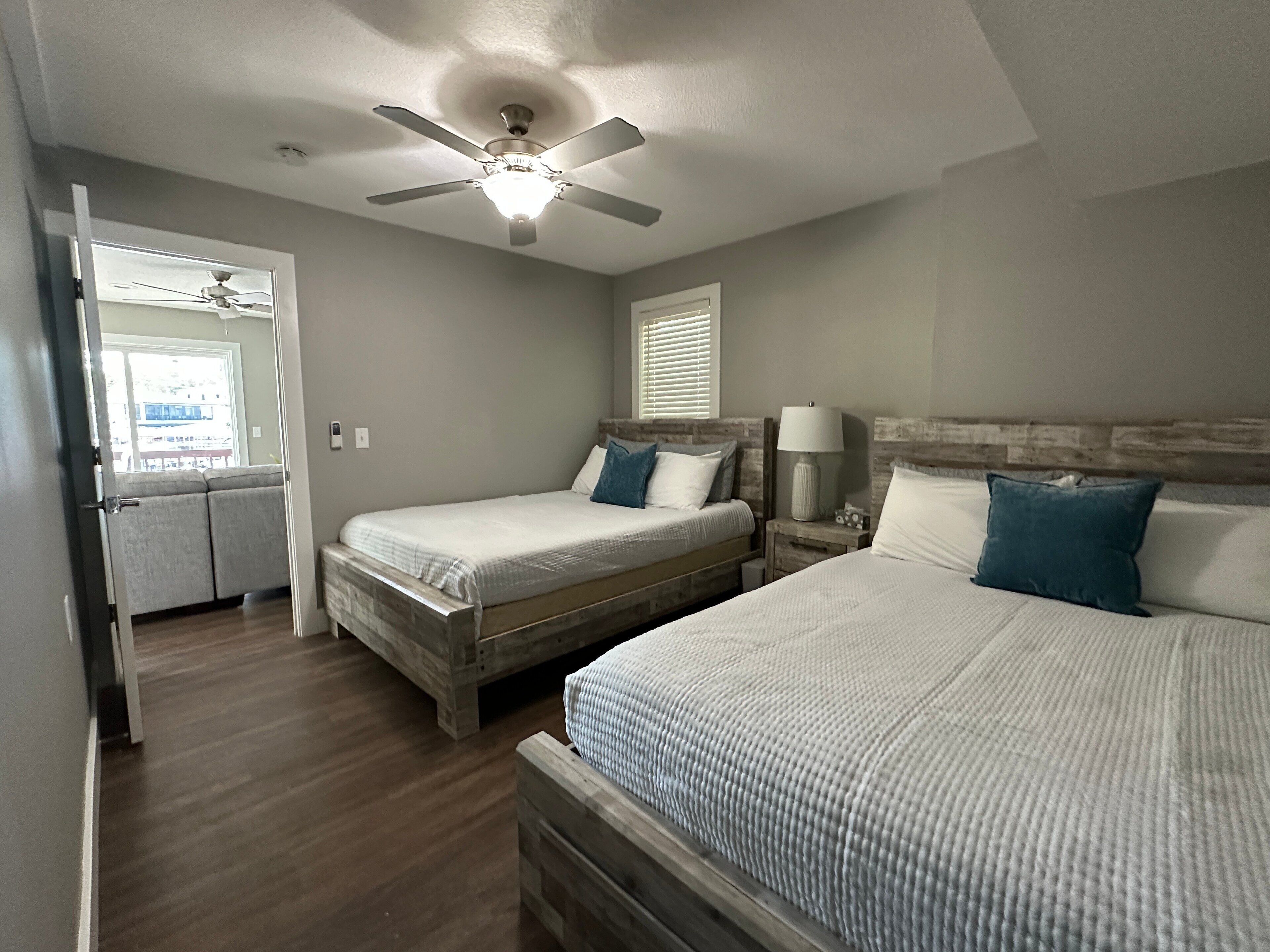 A bedroom with two beds, a ceiling fan, a window with blinds, and a bedside table with a lamp.
