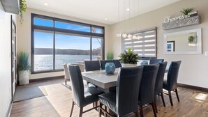 A dining area with a large window overlooking a body of water, a modern chandelier, and a 'Cheers' sign on the wall. - Loto Pointe, Updated Luxury Home w/ Boat & PWC Slip, Boat Ramp, Firepits, Pool Table & More! (Versailles)
