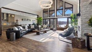 A spacious living room with a large sectional sofa, a coffee table, and a fireplace. There is a kitchen in the background with a stone backsplash and a large window. - Loto Pointe, Updated Luxury Home w/ Boat & PWC Slip, Boat Ramp, Firepits, Pool Table & More! (Versailles)