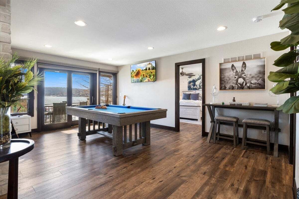 A room with a pool table, a bar area with stools, and a large window offering a view of the outdoors.