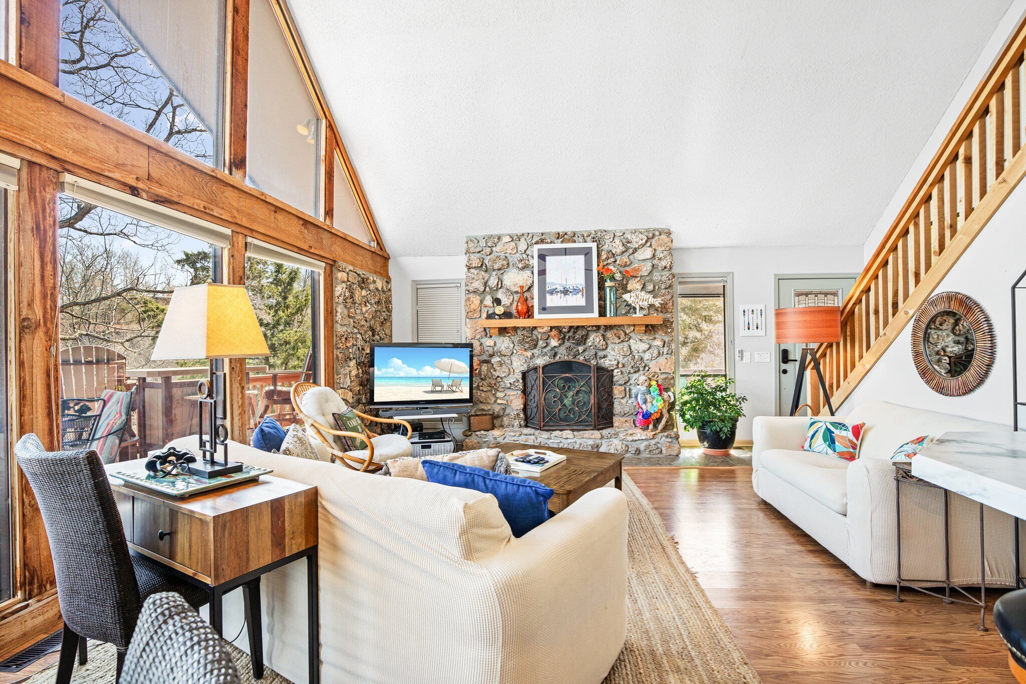A spacious living room with a stone fireplace, wooden beams, and a large window overlooking a deck.