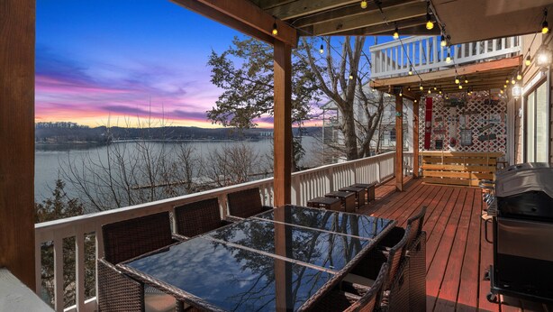 A deck with a glass table and wicker chairs, overlooking a lake with a sunset view. - Amazing Views Close to the Strip - Private Dock! (Lake Ozark)