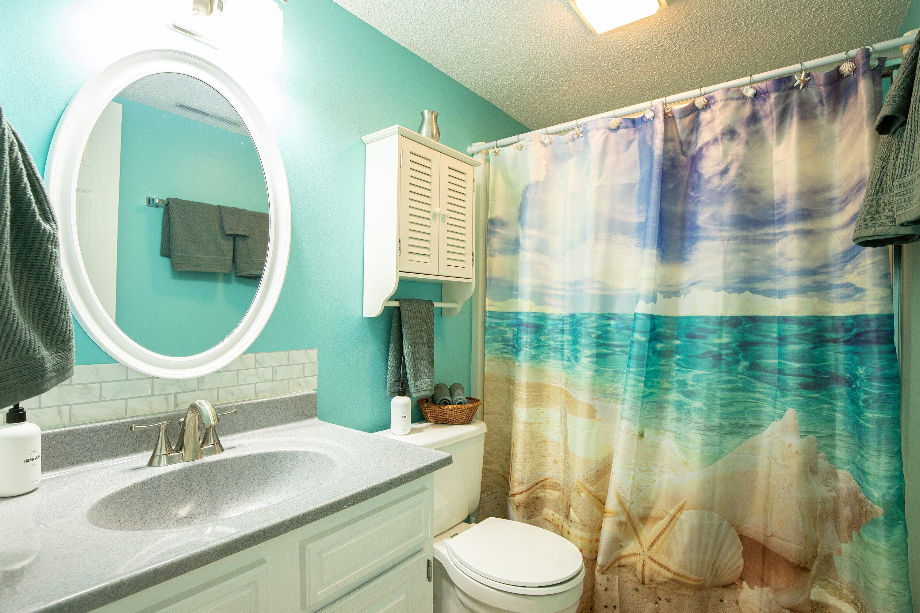 A bathroom with a turquoise wall, a white sink, a mirror, a toilet, and a shower curtain with a sea theme.