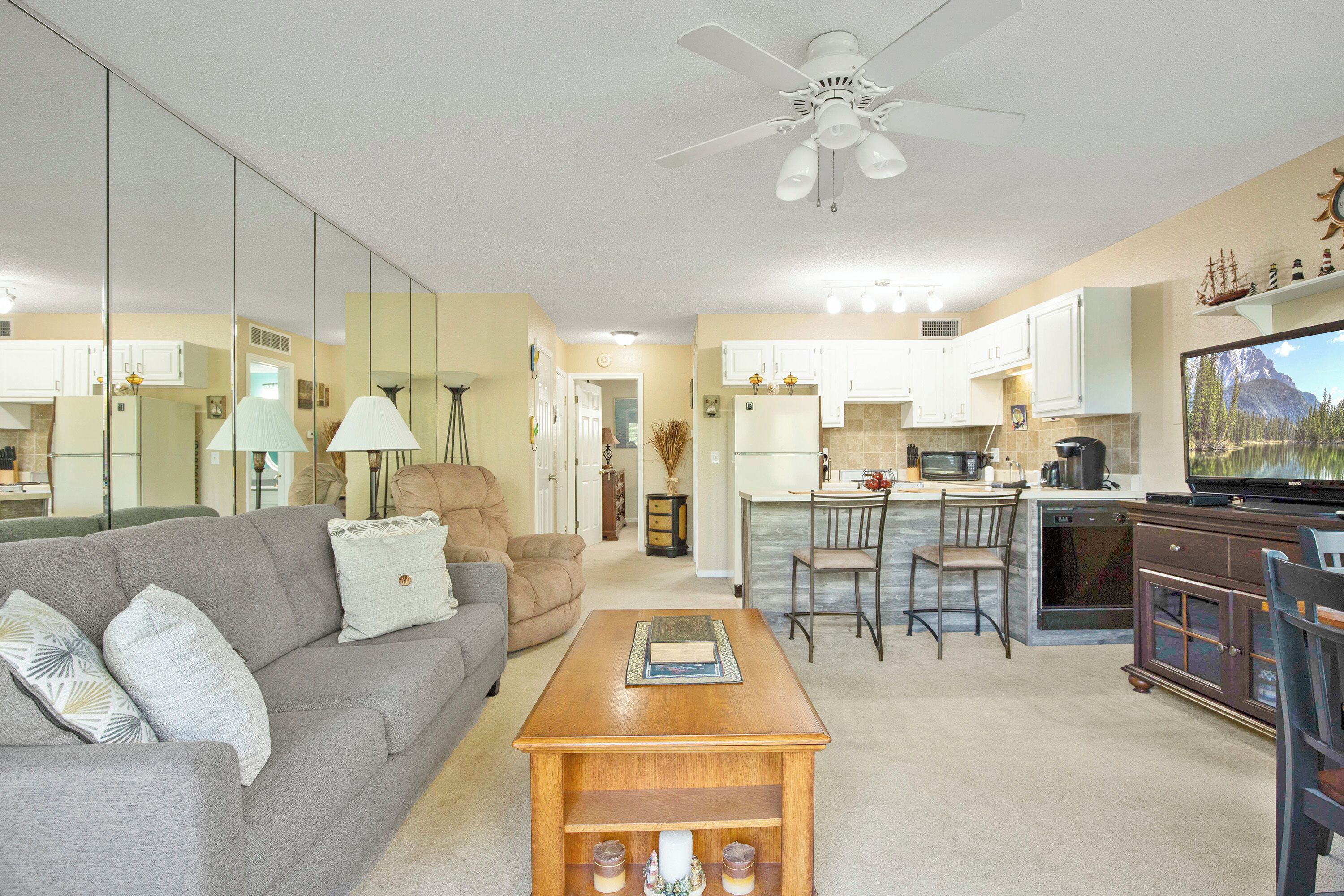 A living room with a sofa, coffee table, and a TV. There is a kitchen area with cabinets and a microwave.