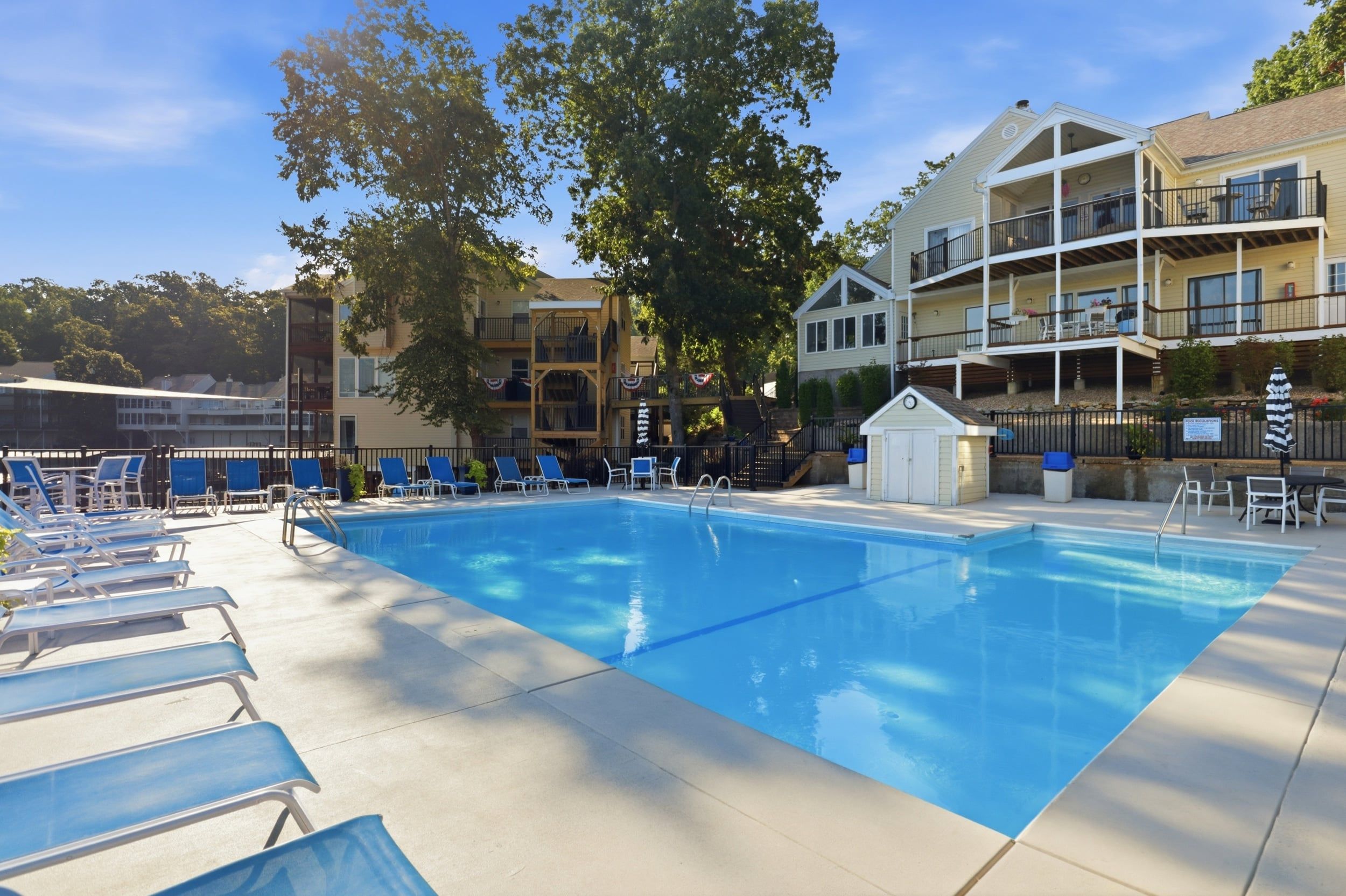 A swimming pool with lounge chairs and a small building, surrounded by trees and a multi-story building with balconies.