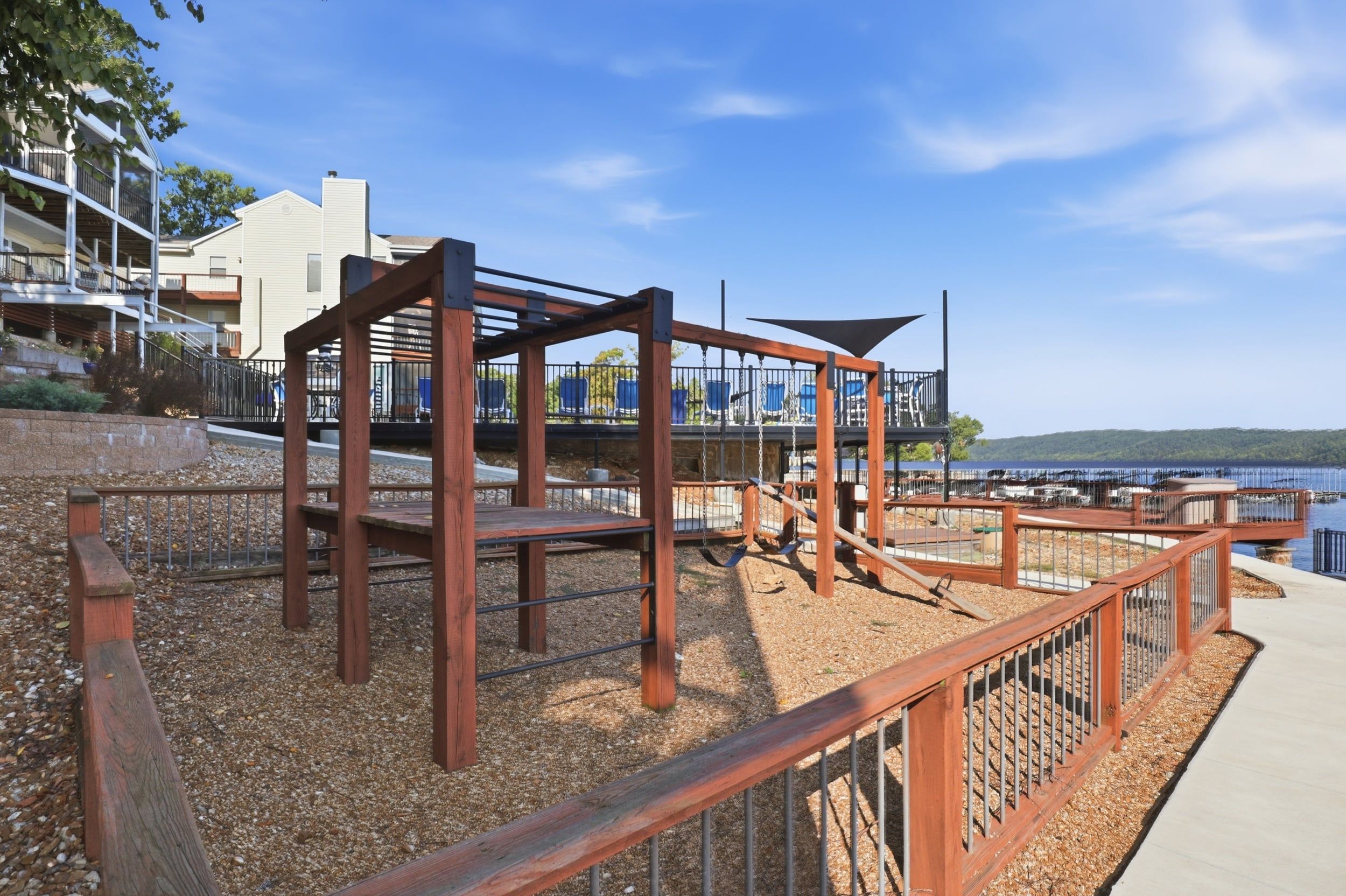 A playground with swings and a climbing structure, surrounded by a wooden fence and a pebble ground.