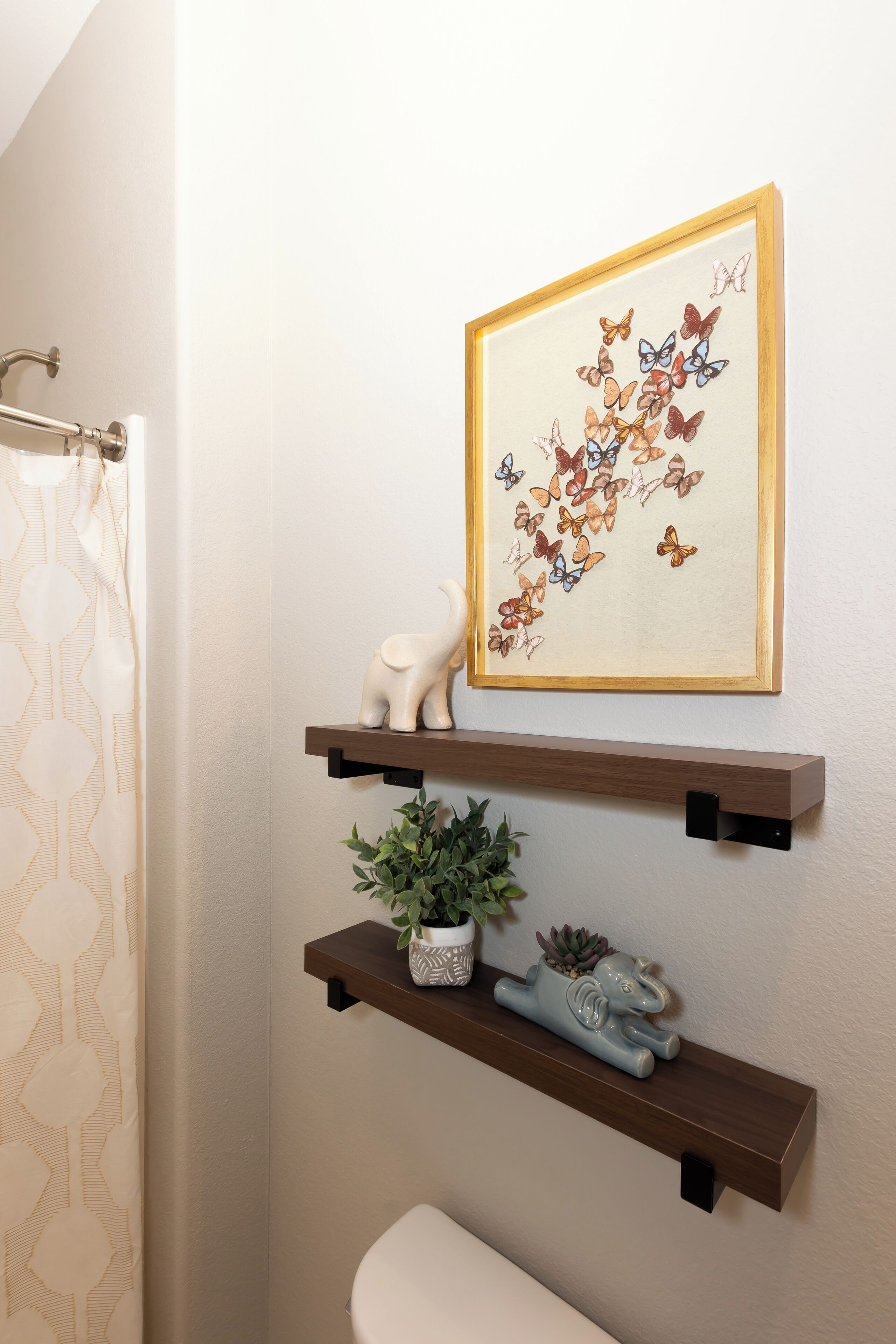 A bathroom with a shower curtain, a framed butterfly artwork, and two shelves with decorative items.