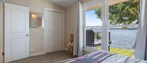 A bedroom with a large window overlooking a body of water, a bed with a striped blanket, and a door with a mirror and light fixture.