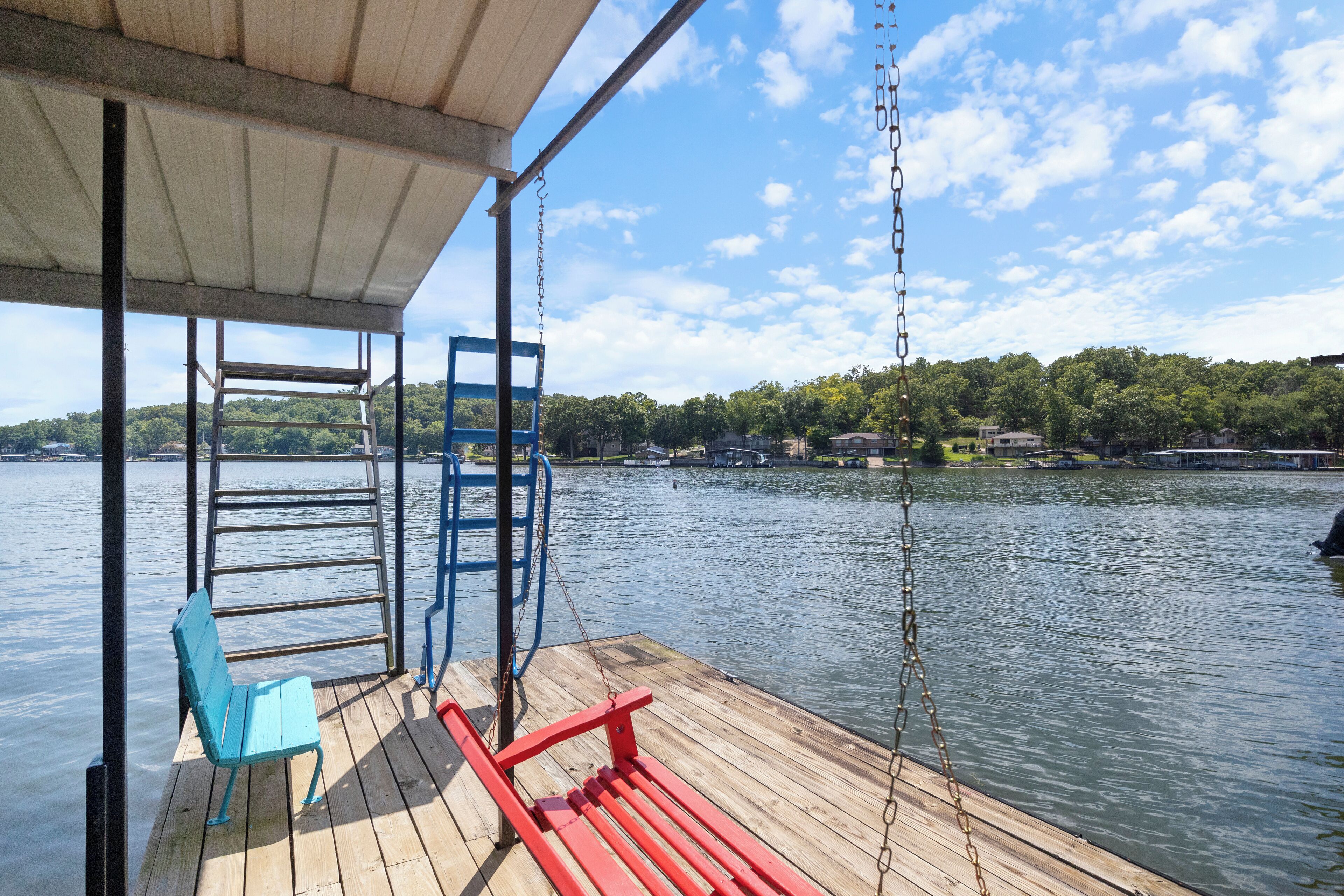 A wooden dock with a blue chair and a red bench, overlooking a calm lake with a forested shoreline.