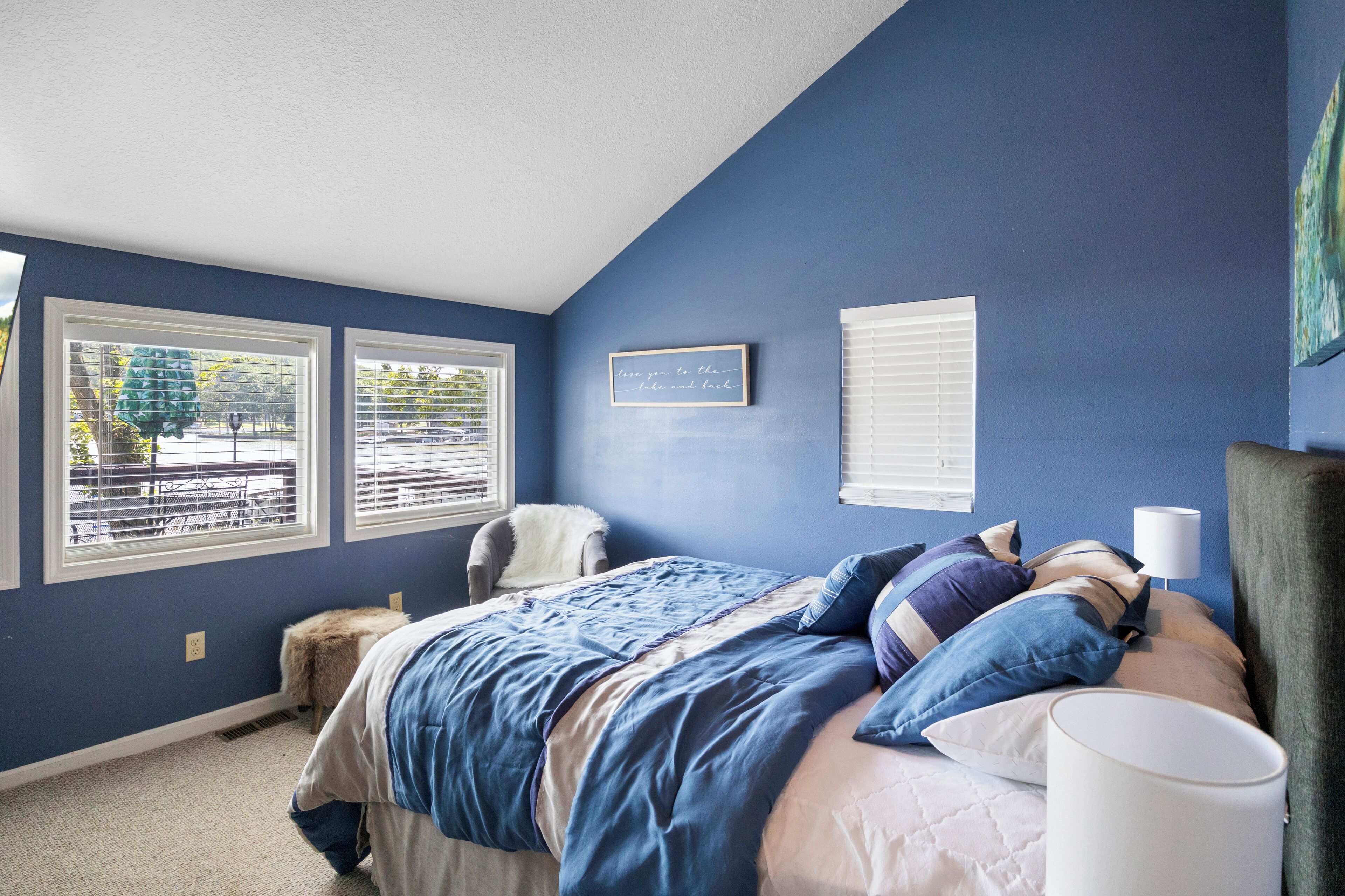 A bedroom with a bed, two windows, a chair, and a wall with a framed quote.