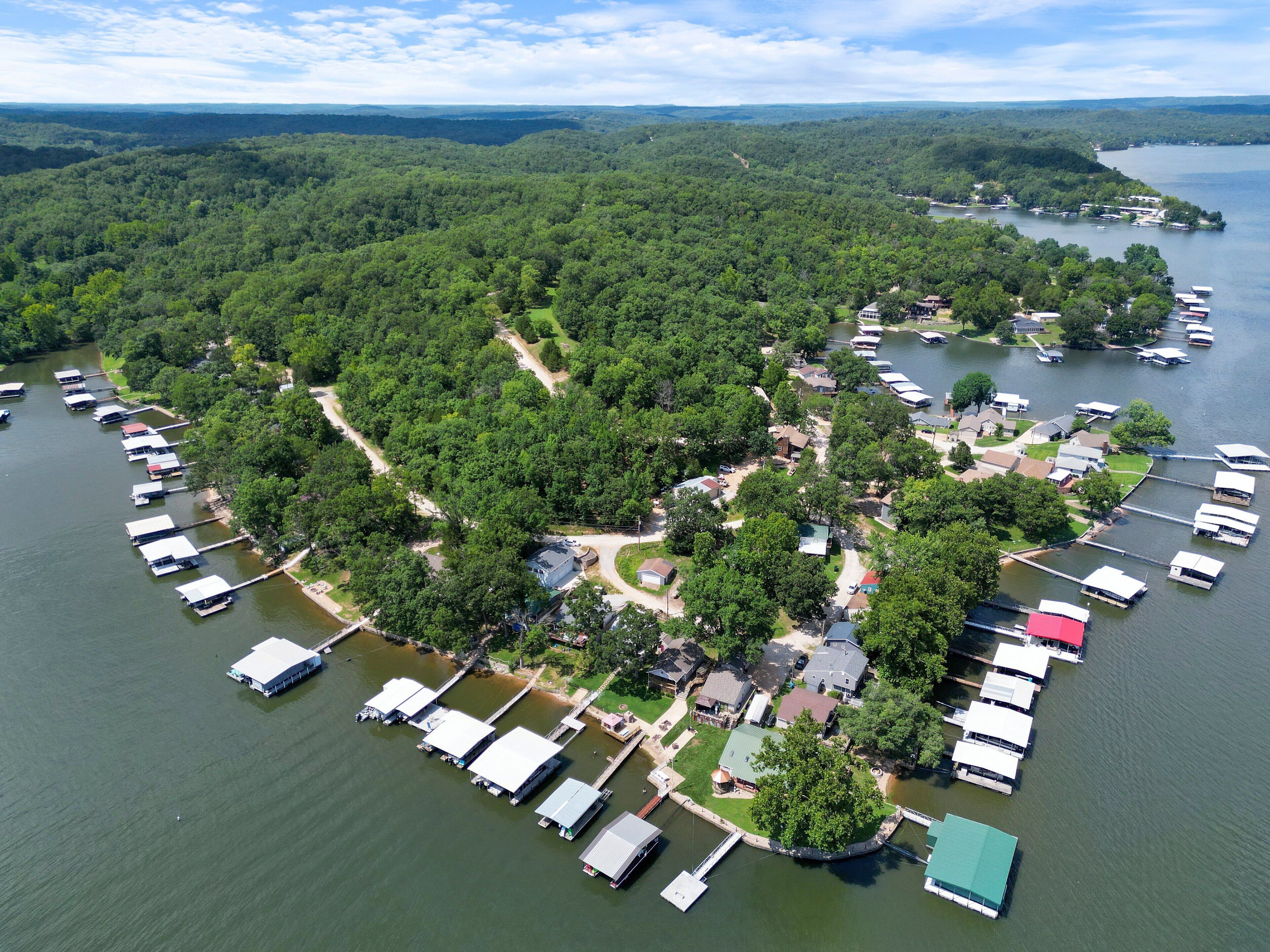 An aerial view of a lakeside property with multiple docked boats, surrounded by dense forest, and featuring a mix of residential and commercial buildings.