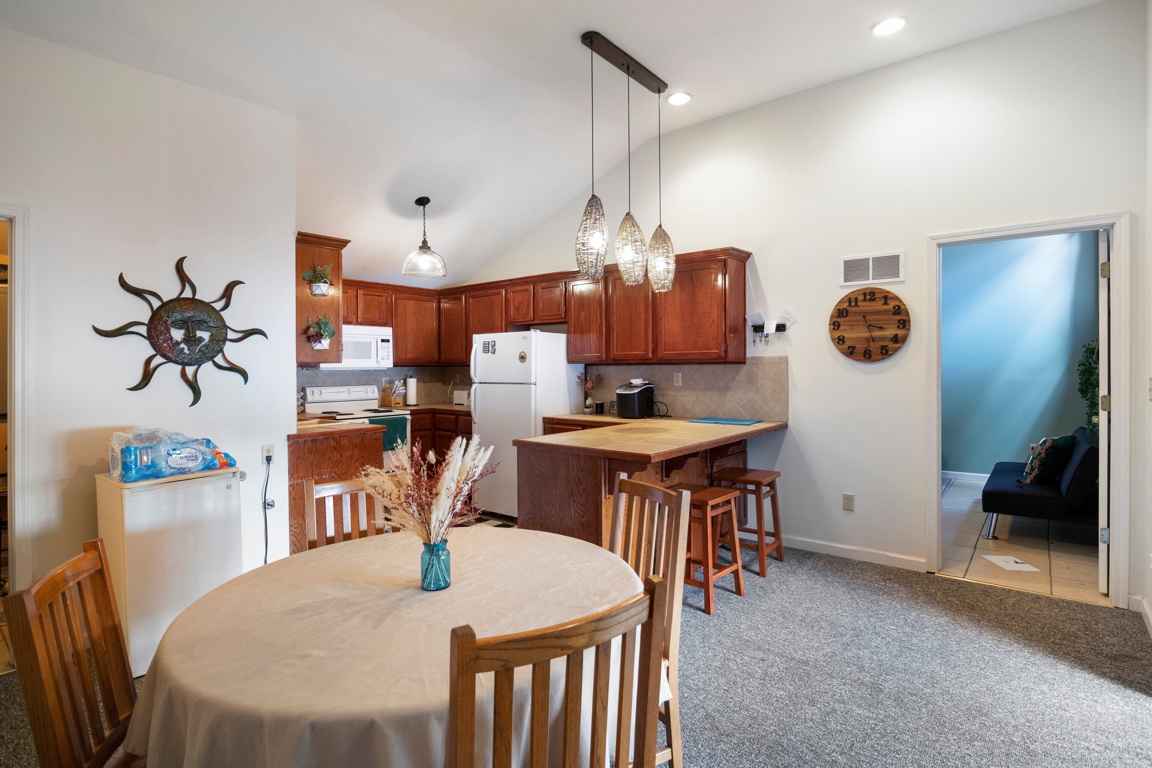 A modern kitchen with wooden cabinets, a central island, pendant lights, and a dining area with a round table and chairs.