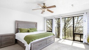 A bedroom with a bed, nightstands, a ceiling fan, and large windows. - Treetop Vibes on Horseshoe Bend (Lake Ozark)