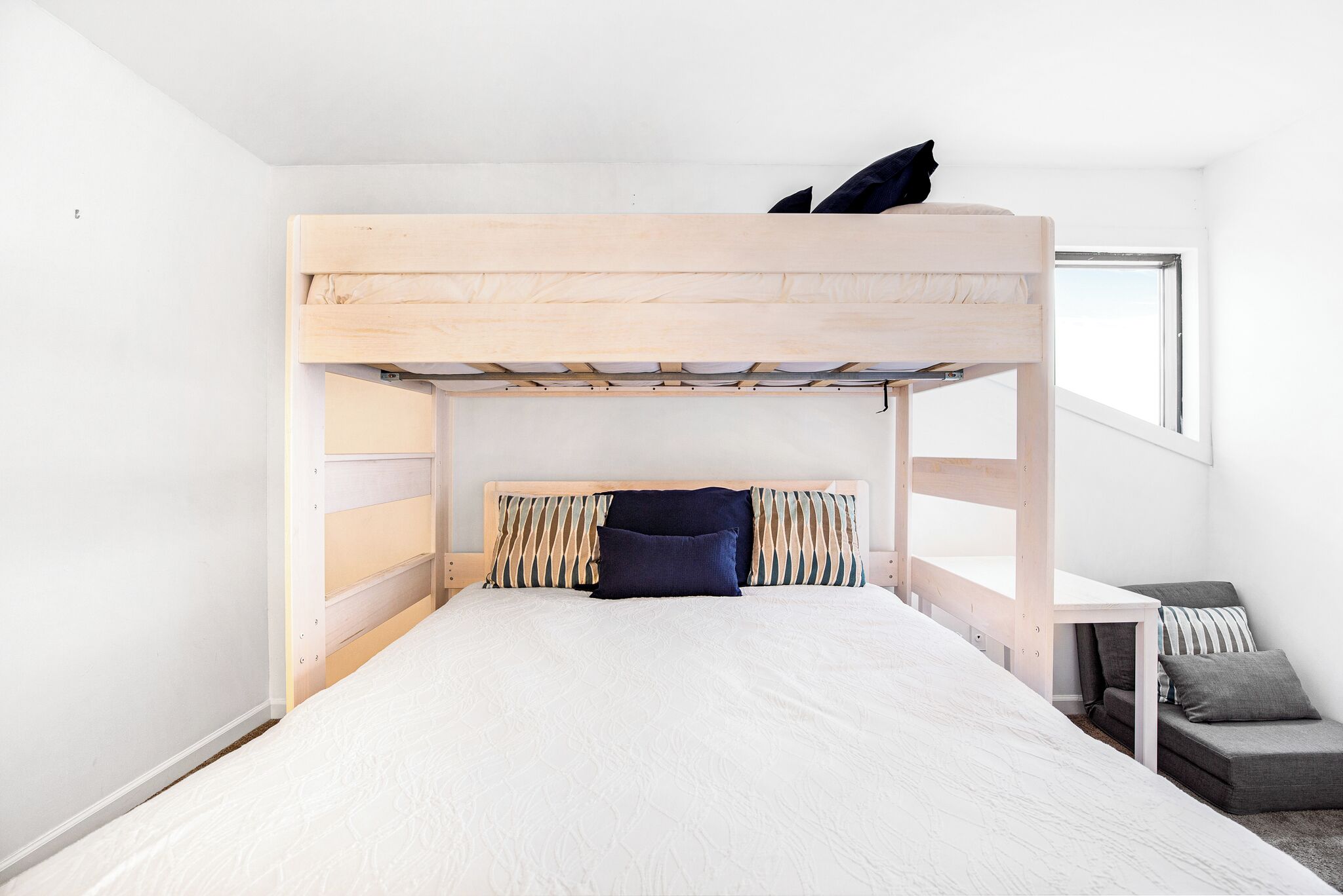 Una cama individual con estructura de madera y colchón blanco. Junto a la cama, hay una mesita con un cojín.