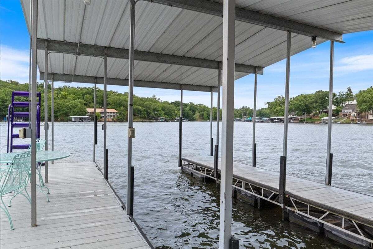 A covered dock with metal chairs and tables, extending over calm water with a forested shoreline in the background.