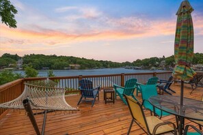 A wooden deck with outdoor furniture, including chairs and a table, overlooking a body of water and a forested area. - Lakefront Fishing Retreat w/ Dock, Hot Tub & Deck Views (Sunrise Beach)