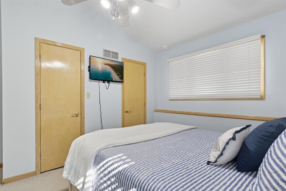 A bedroom with a bed, a TV mounted on the wall, a window with blinds, and a ceiling fan.