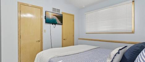 A bedroom with a bed, a TV mounted on the wall, a window with blinds, and a ceiling fan.