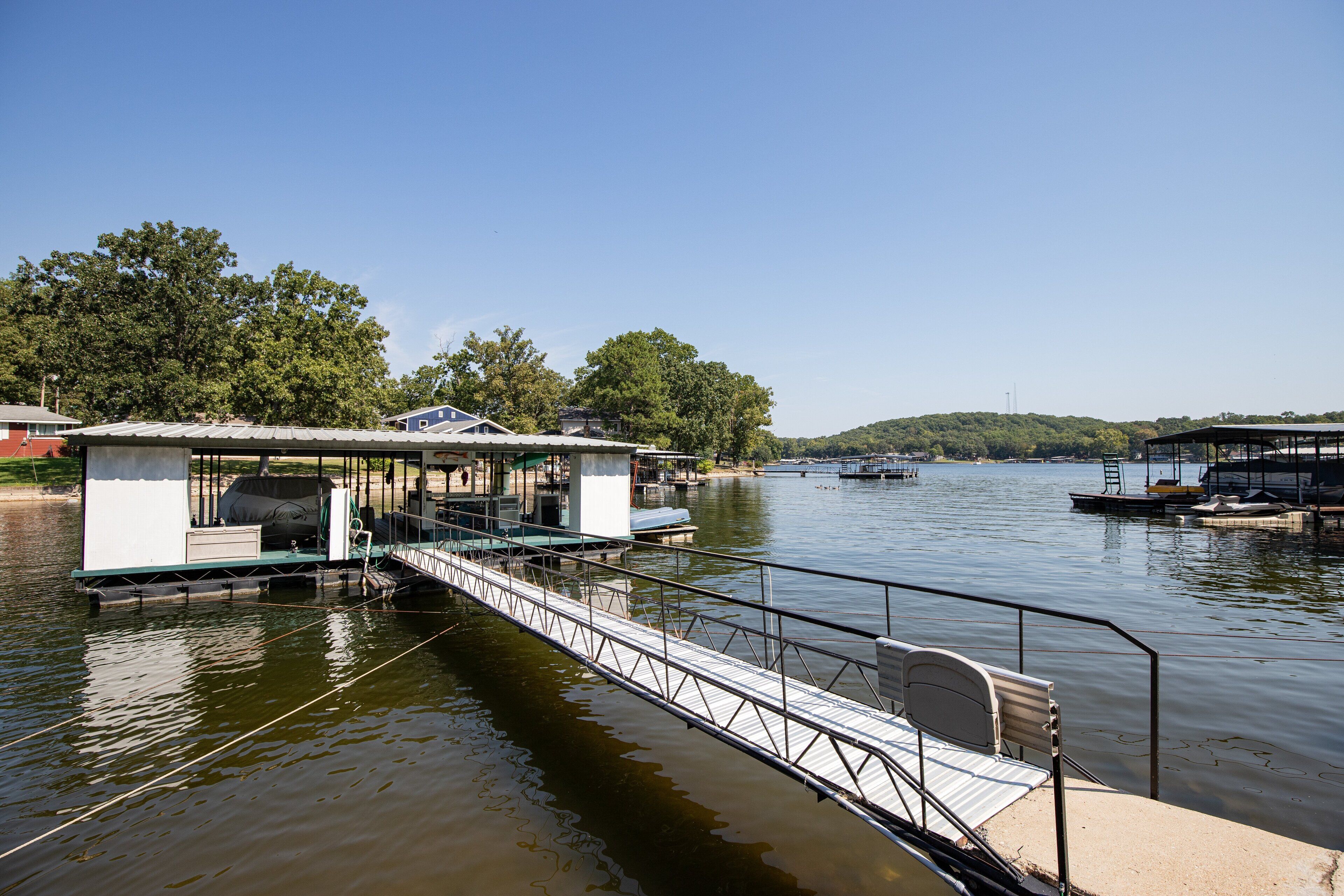 Un muelle con una zona de estar techada y una pequeña construcción, presumiblemente un cobertizo para barcos, junto a un lago tranquilo con barcos y árboles al fondo.