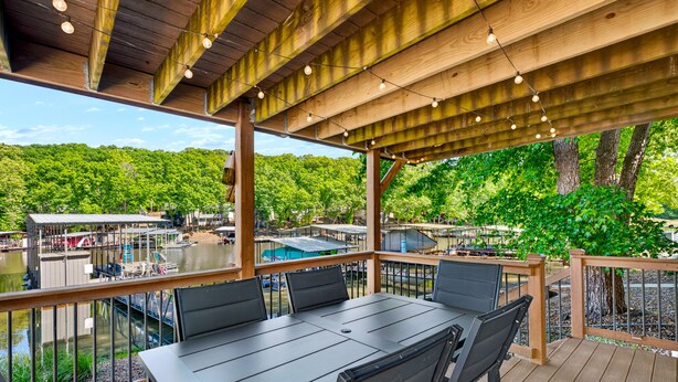 An outdoor deck with a table and chairs, overlooking a body of water and a forested area. - Newly Remodeled w/ Dock, Boat & PWC Slips, Pool Table & Golden Tee (Osage Beach)