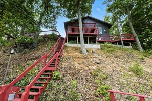 Une cabane avec un escalier rouge menant à un balcon, entourée d’arbres et d’un mur de pierre.