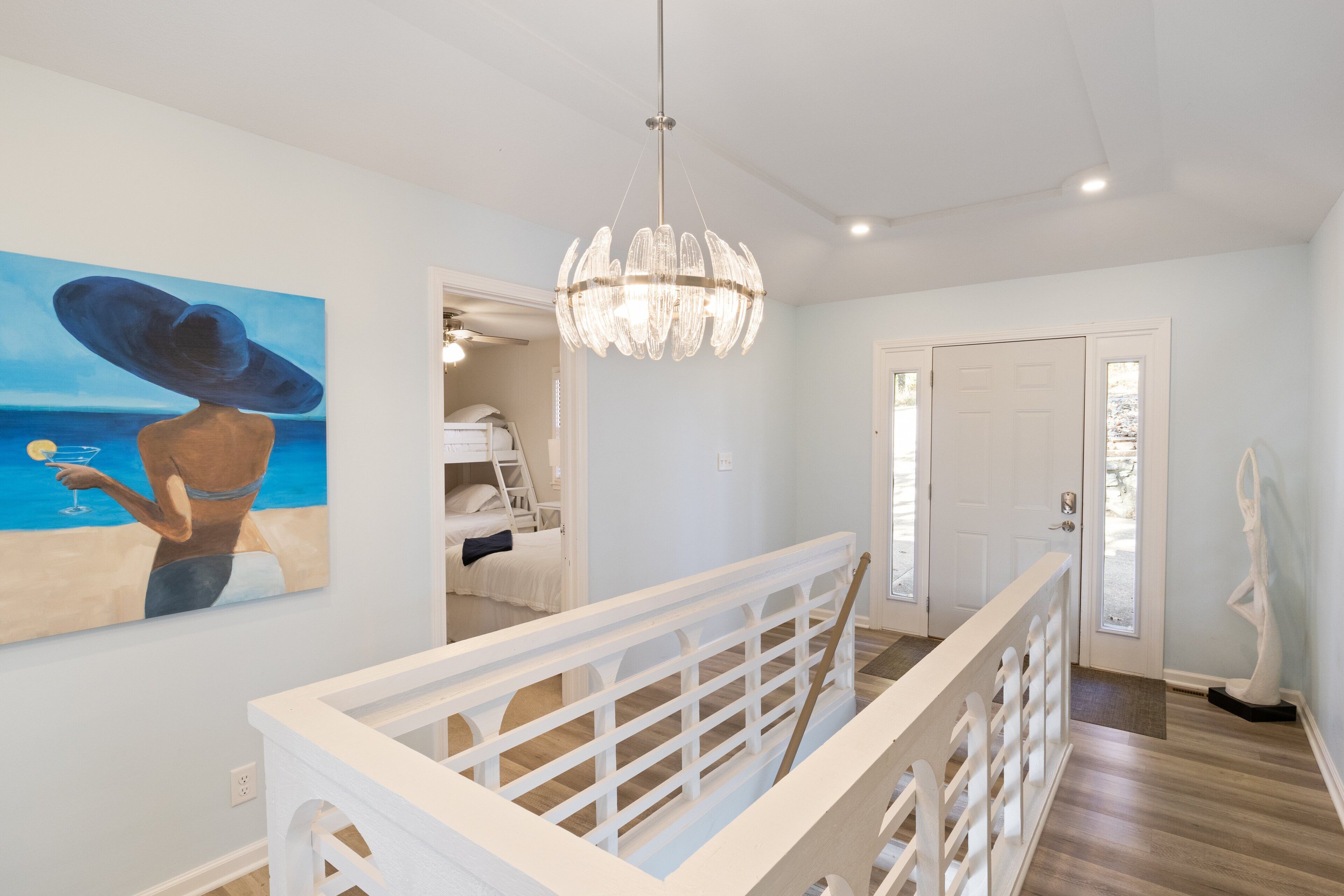 Ein modernes Interieur mit einer Chandelüster, einem Gemälde einer Frau am Meer und einer weißen Geländer.
