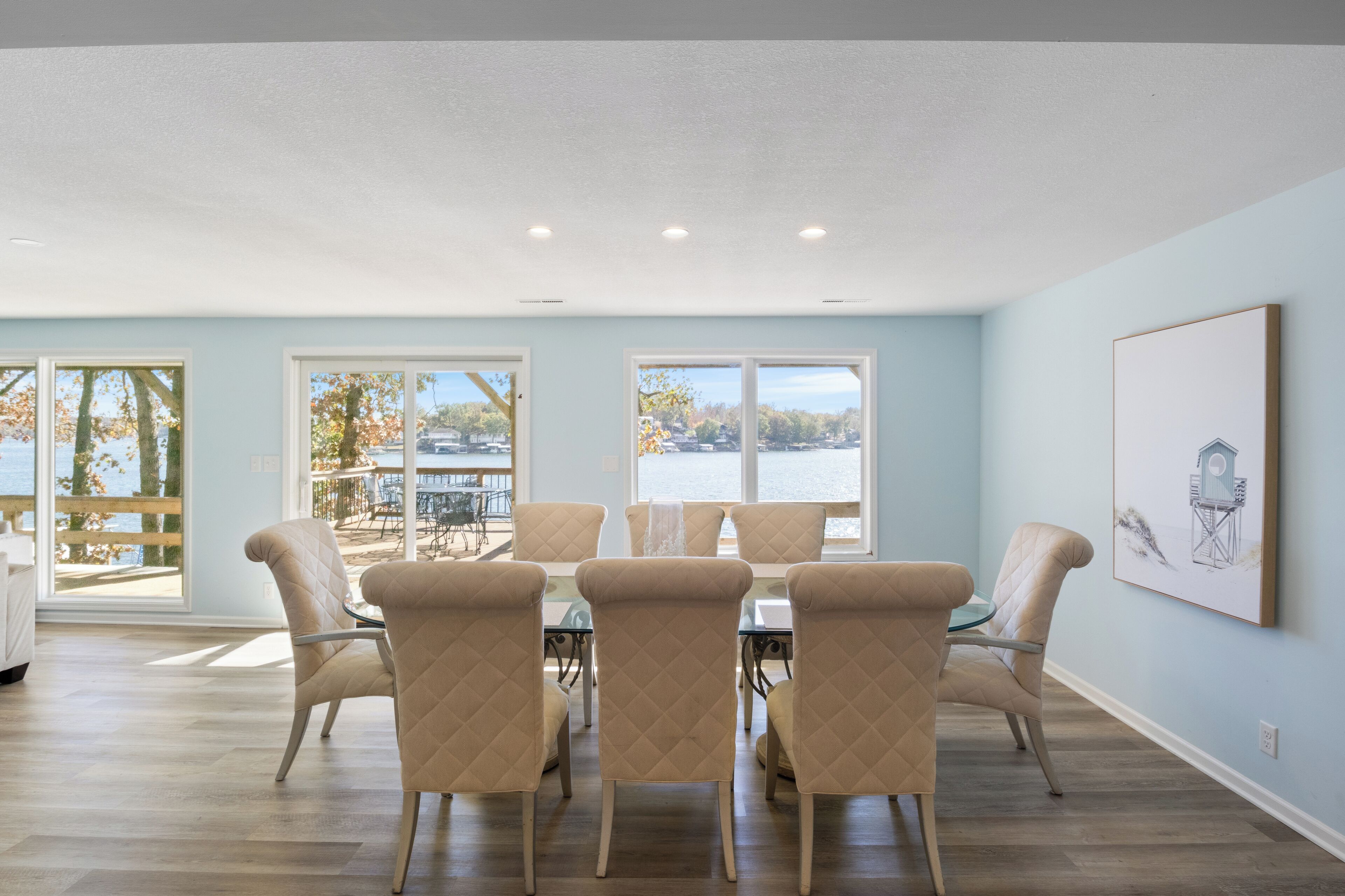 Un comedor con mesa y sillas, ventanas amplias con vista a árboles y un cuerpo de agua, y un cuadro en la pared.