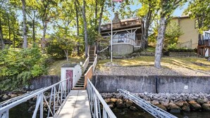 A wooden bridge leading to a house with a deck, surrounded by trees and a body of water.