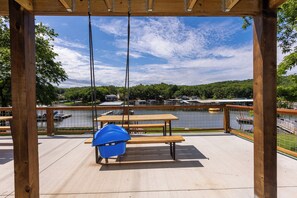 Een houten terras met een picknicktafel en een blauwe emmer, met uitzicht op een meer met boten en een steiger.