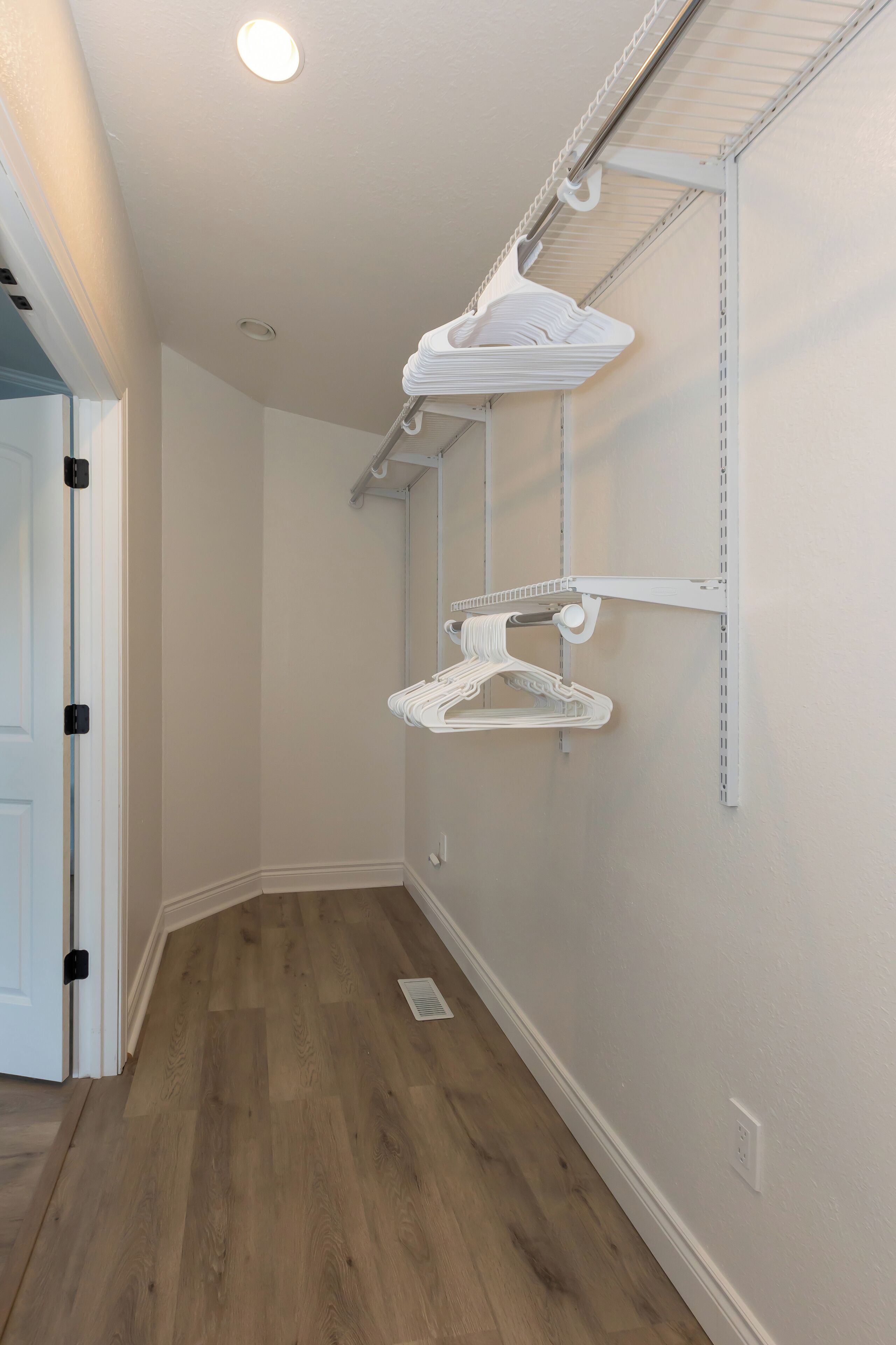 A hallway with wooden flooring, white walls, and a ceiling light. There are white wall-mounted shelves with hangers.