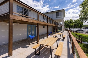 Een terras buiten met houten tafels en banken, een overdekt zitgedeelte met blauwe stoelen, en uitzicht op een gebouw met grote ramen en een witte gevel.