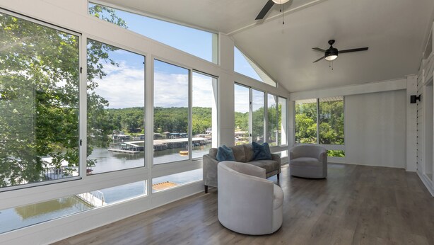 A spacious room with large windows, a ceiling fan, and a view of a lake and forest. - Great Location & Lake Views, Boat Slip (Lake Ozark)