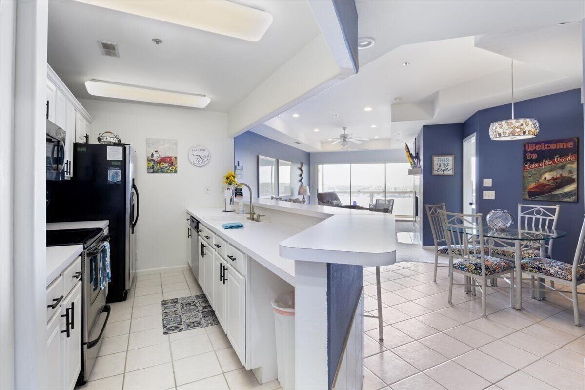 A modern kitchen with white cabinets, a black refrigerator, and a large island with a sink. There is a dining area with a glass table and patterned chairs.