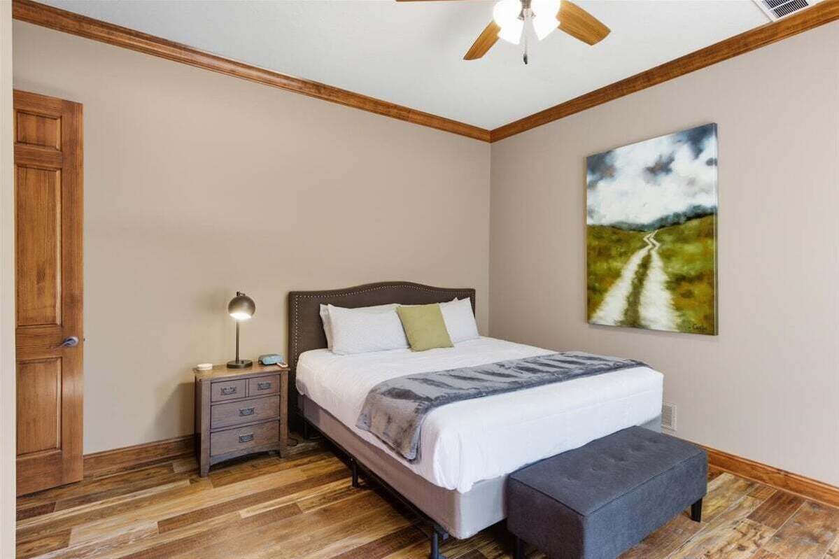 A bedroom with a wooden floor, a bed with a white comforter, a nightstand with a lamp, and a painting of a road on the wall.