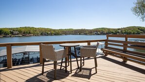 Una terraza de madera con sillas de mimbre y una mesa con vistas a un lago donde se ven barcos y casas al fondo.