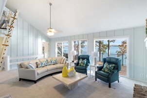 A spacious living room with a curved sofa, a coffee table, and two armchairs. Large windows offer a view of the outdoors.