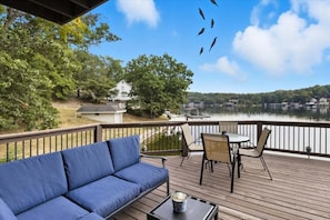 A deck with a blue sofa, a table, and chairs, overlooking a lake with boats and a house in the background. - Expansive Deck, Beautiful Views, Private Dock & Boat Slip (Sunrise Beach)