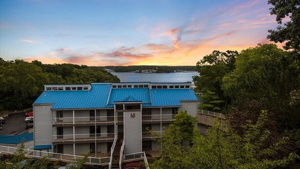 Hotel de vários andares com telhado azul, rodeado por árvores e com vista para um corpo de água.