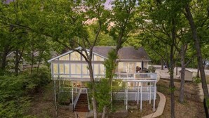 Una casa de dos pisos con porche cubierto y una pequeña terraza, rodeada de árboles y un sendero.