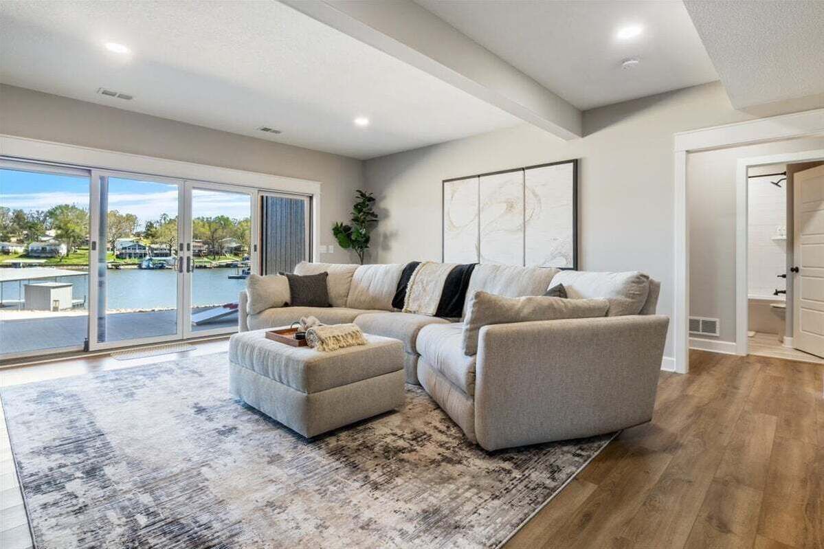 A spacious living room with a sectional sofa, a small ottoman, and a large window offering a view of a parking lot and buildings.