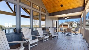 Une terrasse couverte, dotée de fauteuils Adirondack, de grandes fenêtres et offrant une vue sur les maisons au loin.