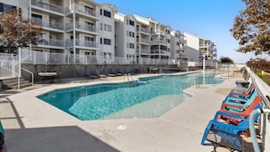 A swimming pool with lounge chairs, a concrete deck, and a multi-story building with balconies.